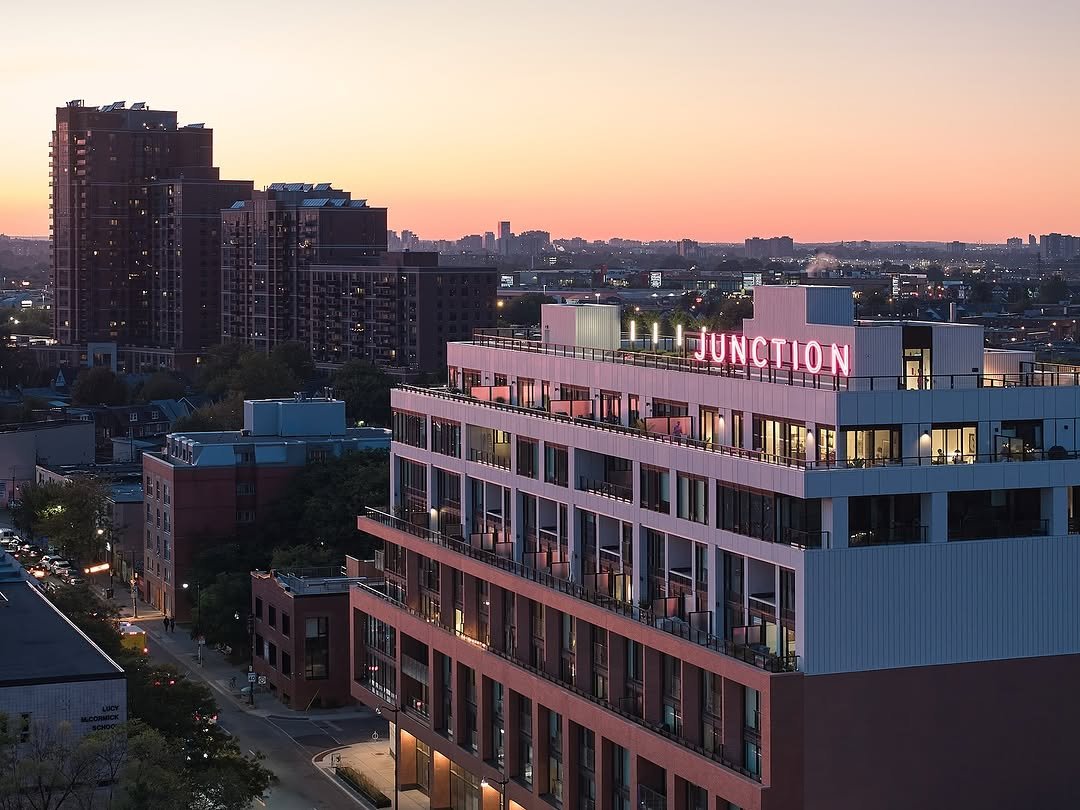 Junction House Condo at 2720 Dundas Street West. A tall modern building with pink neon sign reading 'JUNCTION' on top, in an urban cityscape at sunset with other high-rise buildings and a broad sky.