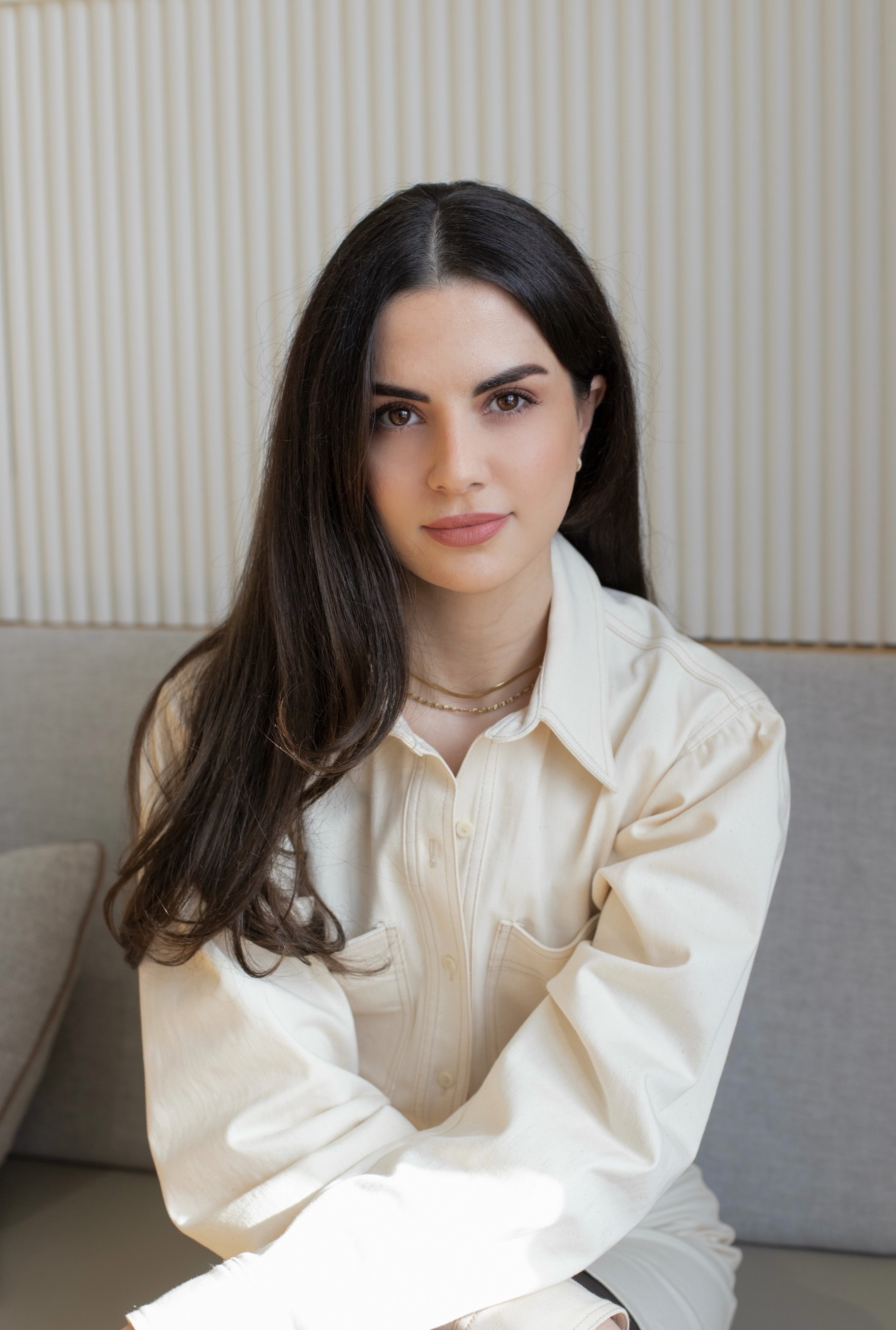 A young woman with long dark hair and brown eyes sitting on a gray sofa in front of a beige curtain, wearing a cream-colored button-up shirt and gold jewelry.