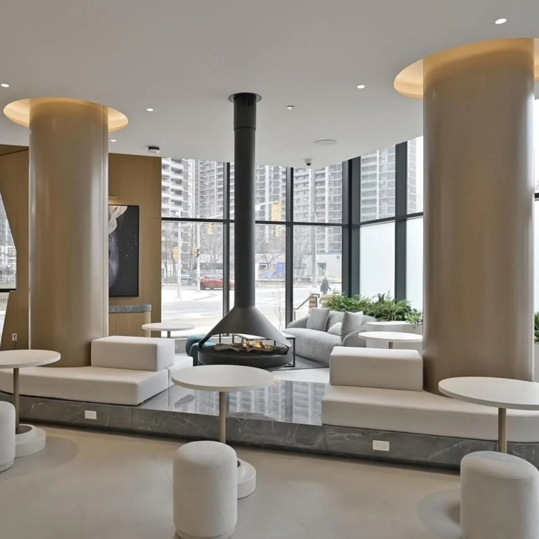 60 Central Park Roadway at Islington Station in Etobicoke Modern lounge area with white couches, round tables, large window, and a central fireplace with black chimney in a high-rise building.